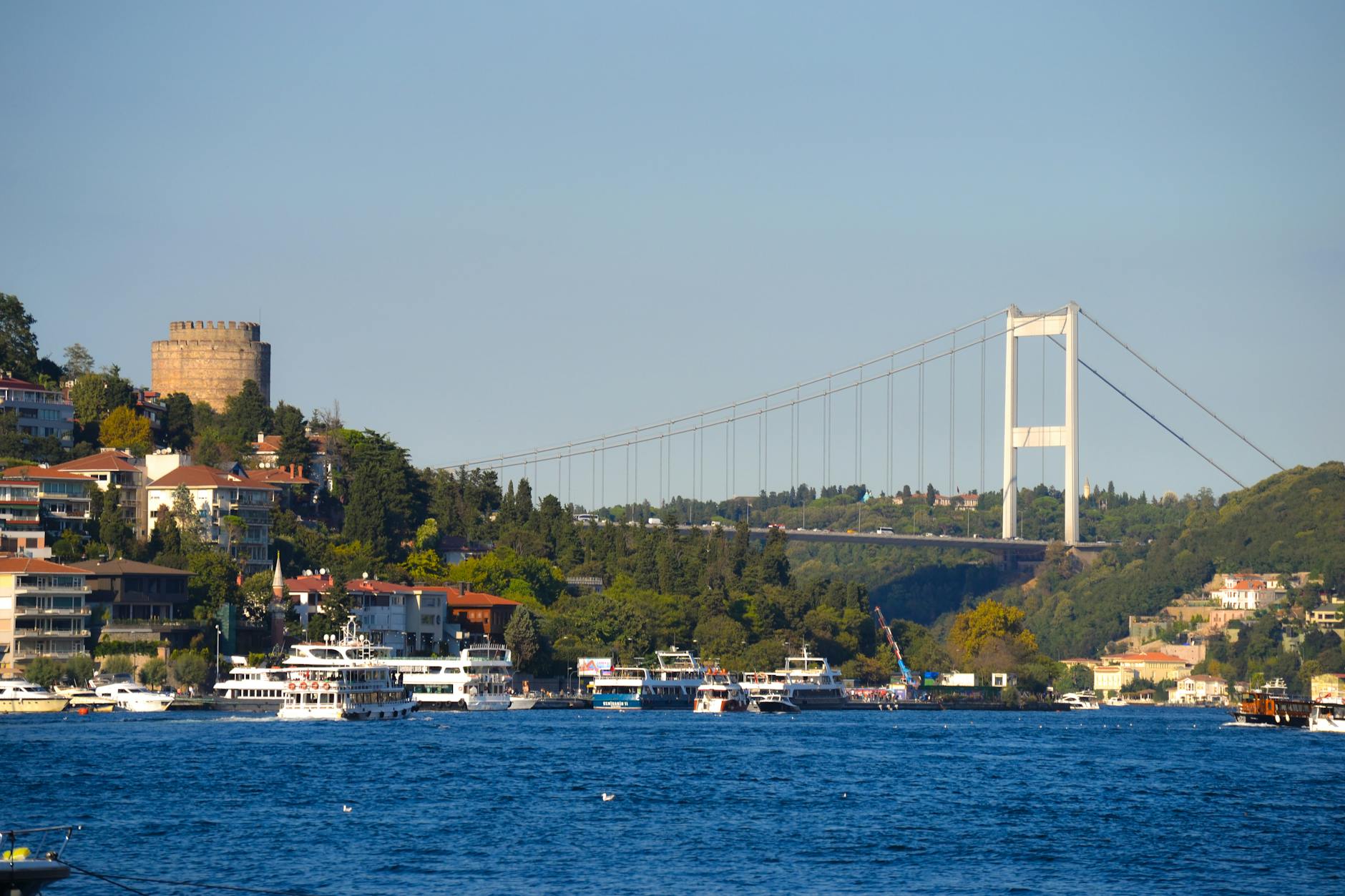 Vista panorámica del Bósforo con el Puente FSM y la Fortaleza de Rumeli Hisarı, con barcos turísticos en primer plano.