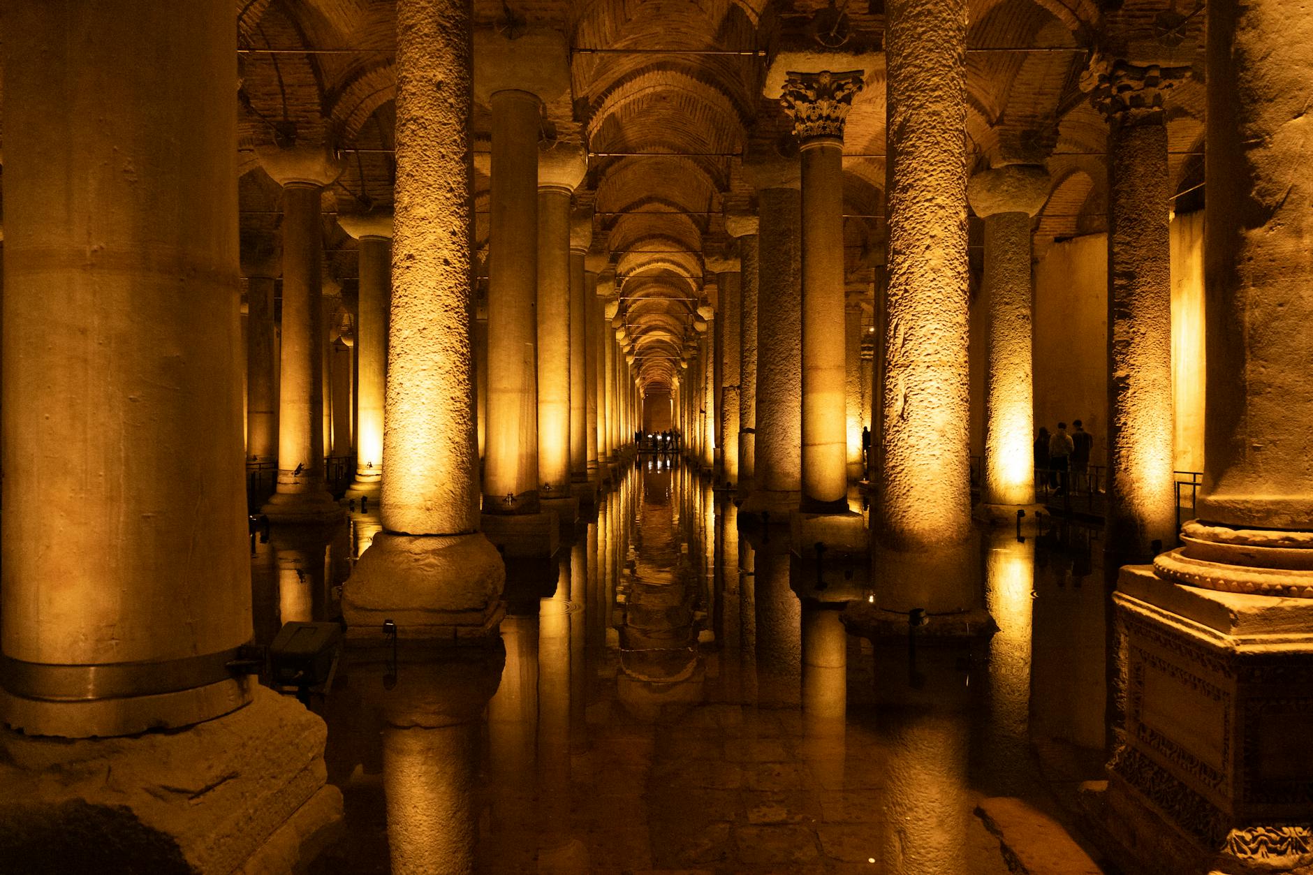 Interior de la Cisterna de Teodosio (Şerefiye) en Estambul, mostrando una larga hilera de columnas antiguas iluminadas con luz cálida y sus reflejos en el agua del suelo.