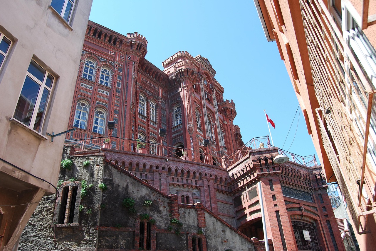 Vista en contrapicado de la fachada de ladrillo rojo del emblemático Colegio Griego Ortodoxo de la Mártir Jorge en Fener, Estambul. Este 'gigante rojo' se alza majestuosamente, con detalles neogóticos y ventanas arqueadas, sugiriendo el esfuerzo que implica subir las cuestas de Fener para llegar a él.