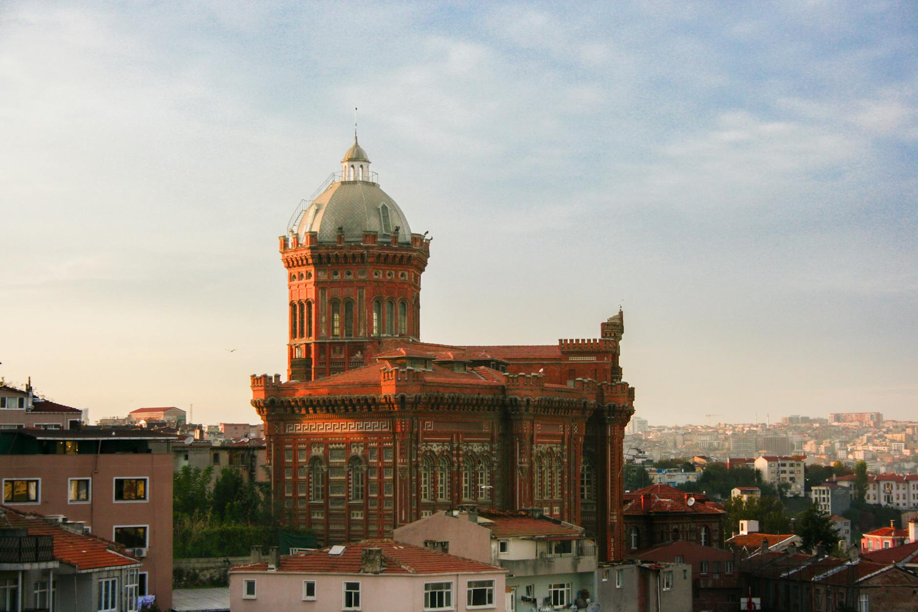 Vista panorámica al amanecer o atardecer del icónico edificio del Colegio Ortodoxo Griego de la Santa Cruz (Fener Rum Erkek Lisesi), un imponente 'gigante rojo' de ladrillo visto con una cúpula plateada, emergiendo sobre los tejados de los barrios de Fener e interior de Estambul. La luz cálida resalta su arquitectura, recordando el esfuerzo de quienes suben "Me dejo los pulmones en las cuestas de Fener por este gigante rojo".