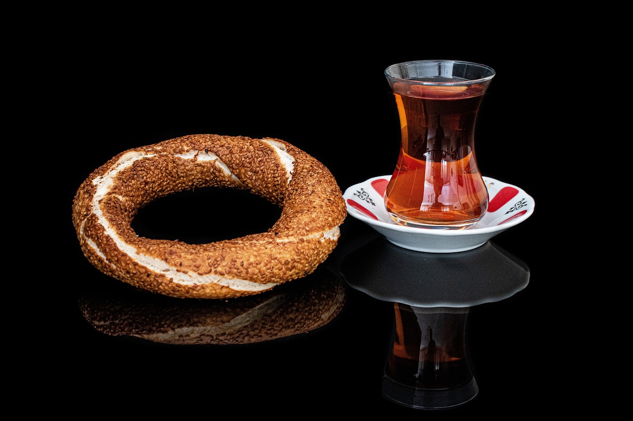 Un pan simit tradicional junto a un vaso de té turco sobre una superficie negra.